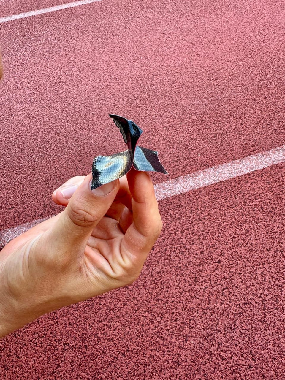 A hand holding a short strip of tape folded into a small stand-up tab, on a running track.
